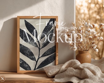 May include: A framed art print featuring a black and white botanical leaf design. The artwork is displayed in a light wood frame. A vase of dried flowers and a folded beige cloth are also in the image. The text "Frame Mockup" is visible.