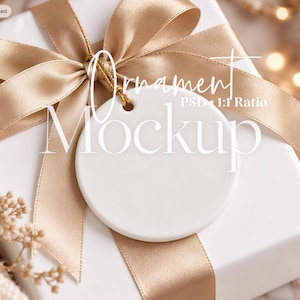 May include: A white circular ceramic ornament with a small hole at the top, attached by a gold cord to a white gift box wrapped in a wide, shiny champagne-colored satin ribbon. The text Ornament Mockup PSD 1:1 Ratio is overlaid on the image.