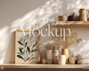 May include: A wooden framed art print featuring a watercolor botanical design with green leaves. The frame is displayed on a wooden shelf with other decorative items, including pottery and dried flowers. The text "Frame Mockup" is visible.