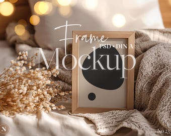 Könnte beinhalten: Ein Holzrahmen-Mockup mit den Wörtern "Frame" und "Mockup" in Weiß. Der Rahmen zeigt ein schwarzes abstraktes Design auf beigem Hintergrund. Das Bild befindet sich auf einem Bett mit einer beigen Decke und getrockneten Blumen.