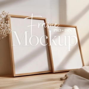 May include: Two wooden picture frames with white inserts are propped against a wall. The frames are leaning on a wooden surface. The image includes dried flowers in a glass vase and the text "Frame Mockup".