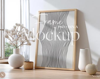 May include: A framed art print with a black and white abstract design, leaning against a white wall. A white vase with a floral arrangement, a small wooden cup, and other decorative vases are on a wooden surface. The text "Frame Mockup" is visible.