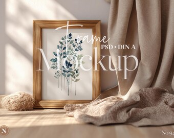 May include: A wooden framed print featuring a watercolor botanical design in shades of blue and green. The print is propped against a wall, with a neutral-colored fabric draped nearby. The word "Mockup" is visible on the print.