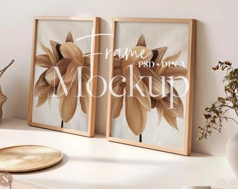 May include: Two vertical light wood frames containing identical prints of a large, stylised beige and brown flower. The text "Frame Mockup" and "PSD DIN A" is overlaid in white script and sans-serif font across the centre of the two frames.
