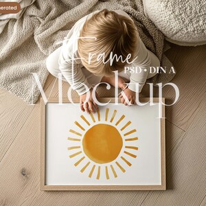 May include: A framed art print with a watercolour sun design, yellow centre and radiating yellow lines. The artwork is in a light wood frame. The image is on a light wood floor with a child in the background.