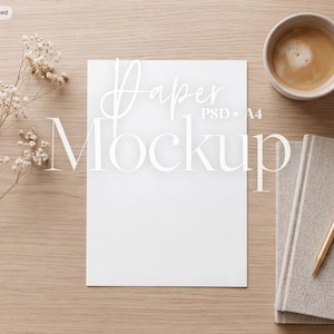 May include: A blank white A4 paper mockup centered on a light wood surface, accompanied by a sprig of dried white flowers, a ceramic mug filled with coffee, a grey notebook, and a gold pen. The text Paper Mockup PSD A4 is visible.