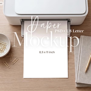 以下が含まれることがあります： 白いプリンターから出力された白い紙には「Paper Mockup PSD US Letter 8.5 x 11 inch」の文字が印刷されています。横には金色のペーパークリップが入った白いボウル、金色のペン、2冊の灰色のノートが明るい木目の表面に置かれています。