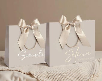 May include: Two light beige gift bags with twisted rope handles and large satin bows. The left bag features the name Samuele in white script. The right bag features the name Selina in white script with the word BRIDESMAID printed below it.
