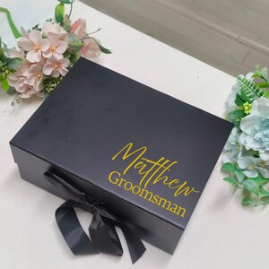 May include: A rectangular black gift box featuring a black ribbon tied in a bow on the front. The lid is personalized with the name Matthew and the word Groomsman printed in a gold script and sans-serif font combination.