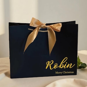May include: A navy blue gift bag with a gold satin bow and rope handles. The bag has the name "Robin" in gold script, with "Merry Christmas" below. A festive holiday gift bag.