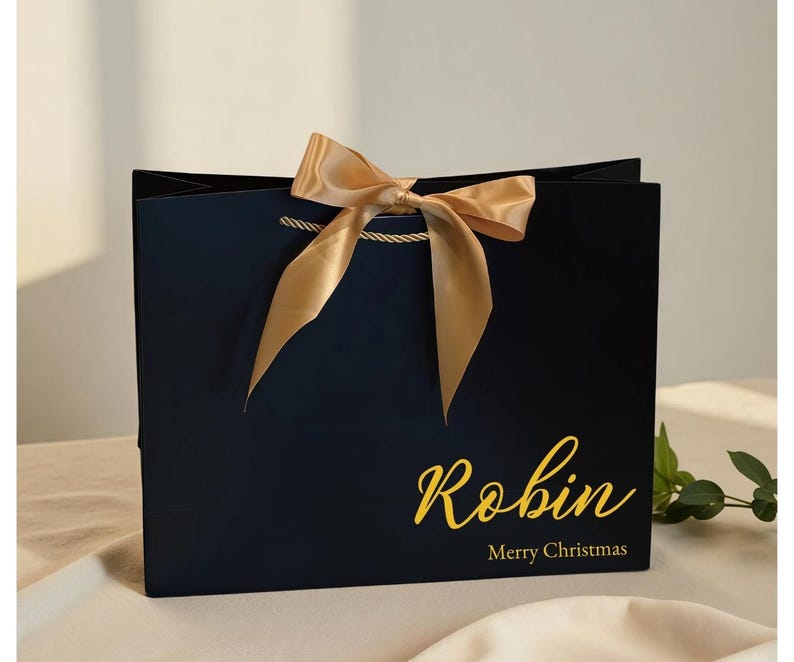 May include: A navy blue gift bag with a gold satin bow and rope handles. The bag has the name "Robin" in gold script, with "Merry Christmas" below. A festive holiday gift bag.