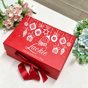 May include: A rectangular red gift box featuring a white graphic print of hanging Christmas ornaments, a snowman, and the text "Lackie Christmas Eve Box" in white script. The box is closed with a red ribbon tied in a bow at the front centre.