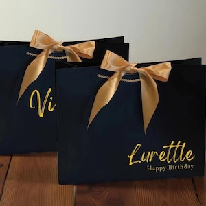 Peut inclure: Deux sacs cadeaux bleu marine avec des nœuds en satin doré et des poignées. L'un porte le nom "Vi" en écriture dorée, et l'autre dit "Lurette Happy Birthday" en écriture dorée. Les sacs sont sur un fond clair.