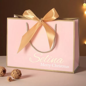 May include: A pink gift bag with a gold bow and handles. The bag has gold trim and the name "Selina" and the words "Merry Christmas" printed on the front. Decorative spheres are in the foreground.