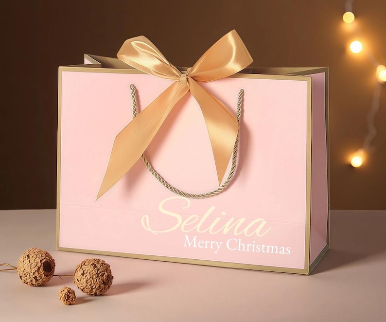 May include: A pink gift bag with a gold bow and handles. The bag has gold trim and the name "Selina" and the words "Merry Christmas" printed on the front. Decorative spheres are in the foreground.