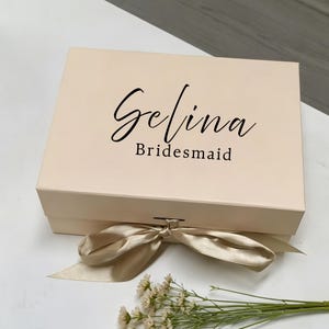 May include: A rectangular, light beige gift box with a hinged lid, featuring the name Gelina in a black script font and the word Bridesmaid in a black sans-serif font. A wide, champagne-colored satin ribbon is tied in a bow at the front.