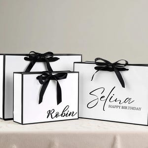 May include: Three white gift bags with black trim and black satin ribbon bows. The bags have the names "Robin" and "Selina Happy Birthday" printed in black script. The bags are on a white surface.