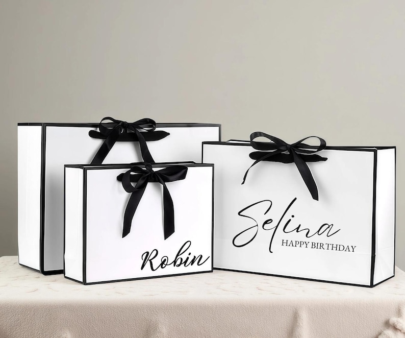 May include: Three white gift bags with black trim and black satin ribbon bows. The bags have the names "Robin" and "Selina Happy Birthday" printed in black script. The bags are on a white surface.