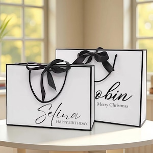 May include: Two white gift bags with black trim and handles, each adorned with a black bow. One bag says "Selina Happy Birthday" and the other says "Robin Merry Christmas" in elegant script.