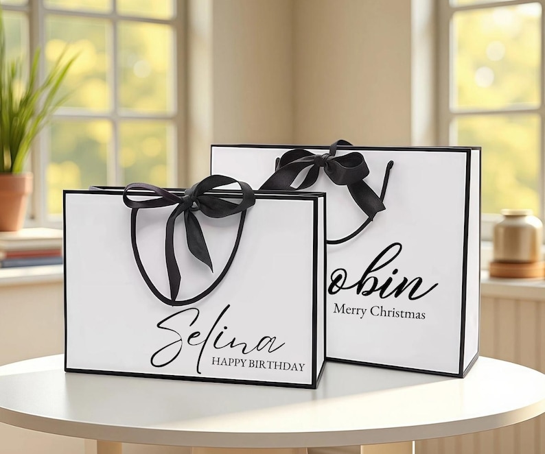 May include: Two white gift bags with black trim and handles, each adorned with a black bow. One bag says "Selina Happy Birthday" and the other says "Robin Merry Christmas" in elegant script.
