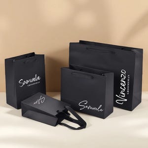 May include: Several black gift bags of different sizes, each with white cursive text bearing names such as "Samuele" and "Vincenzo GROOMSMAN". The bags have black handles and a glossy finish, ideal for presenting gifts.