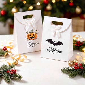 May include: Two white paper gift bags with die-cut handles and white satin bows. One bag features an orange jack-o'-lantern graphic and the name Megan in black script. The other bag displays a black bat graphic and the name Kristen in black script.