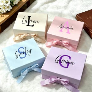 May include: Four square gift boxes with lids and satin ribbons. The cream box features the name Lauren with a large L. The pink box displays the name Amanda with a large A. The light blue box shows the name Shirley with a large S. The lavender box features the name Gemma with a large G.