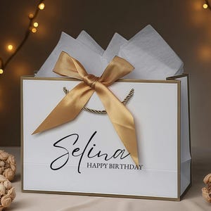 May include: White gift bag with gold trim, a gold satin bow, and twisted gold handles. The bag is filled with white tissue paper and has the name "Selina" and "Happy Birthday" printed on the front.