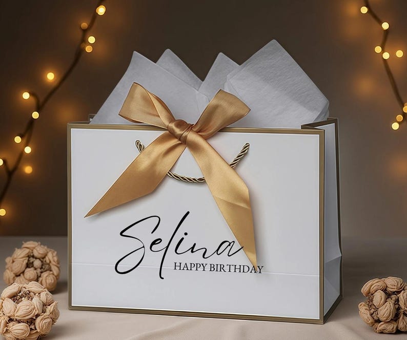 May include: White gift bag with gold trim, a gold satin bow, and twisted gold handles. The bag is filled with white tissue paper and has the name "Selina" and "Happy Birthday" printed on the front.