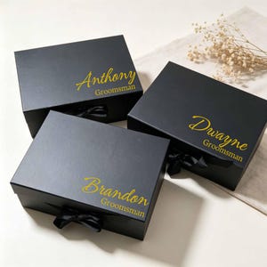 May include: Three rectangular black gift boxes with lids, each featuring a black ribbon bow at the front. The lids are personalized in gold script with the names Anthony, Dwayne, and Brandon, followed by the word Groomsman in print.