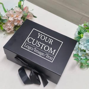 May include: A rectangular black gift box featuring a white rectangular border and the white text "YOUR CUSTOM Logo/Image/Text" printed on the lid. The box is accented with a black ribbon tied in a bow at the front corner of the lid.