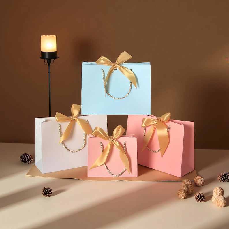 May include: Four gift bags in pastel colors: light blue, white, and pink. Each bag has a gold satin ribbon tied in a bow and a twisted rope handle. The bags are arranged on a light brown surface with decorative pinecones and a candle holder.