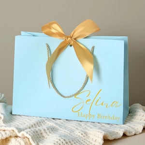 Pode incluir: Uma sacola de presente azul claro com um laço de fita dourada e alças de corda. A sacola tem o nome "Selina" e as palavras "Happy Birthday" impressas em ouro.