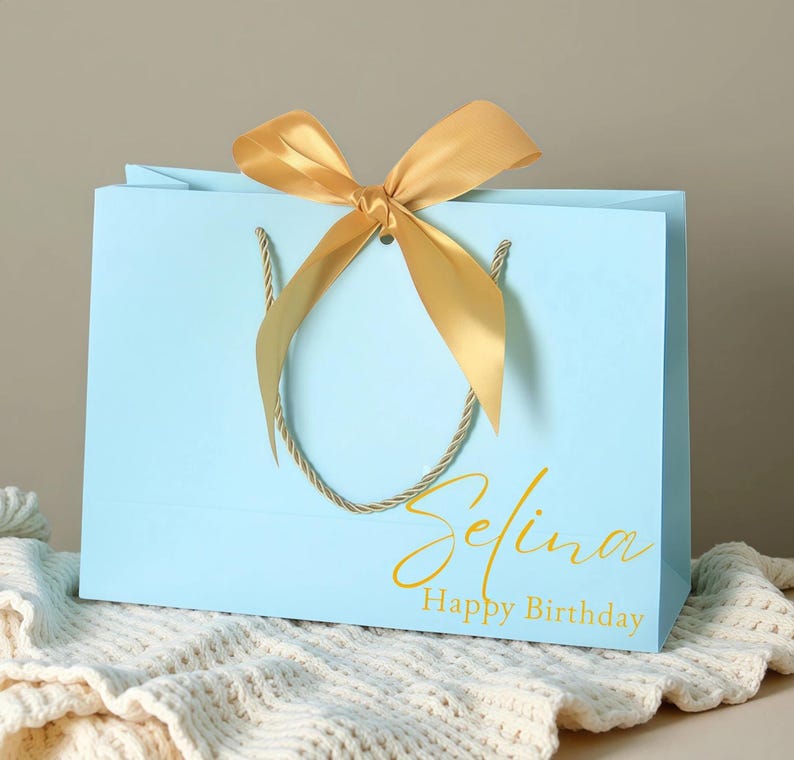 May include: A light blue gift bag with a gold satin bow and rope handles. The bag has the name "Selina" and the words "Happy Birthday" printed in gold script.