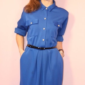 Puede incluir: Un vestido azul real con cuello, mangas largas y cinturón negro. El vestido tiene dos bolsillos en la parte delantera y llega a la altura de la rodilla.