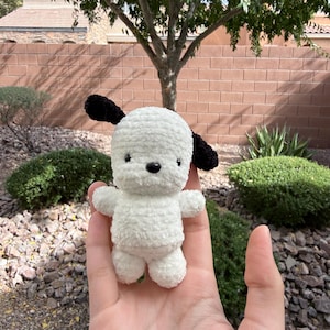 May include: A handmade white and black crochet puppy doll. The plush toy has a fluffy white body, black ears, and a black nose. The doll is small and held in a person's hand, with a blurred background of greenery and a brick wall.