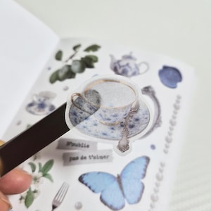 May include: A sticker depicting a teacup and saucer with a blue and gold floral pattern. The sticker is set against a white backdrop with other stickers, including a butterfly and the text "N'oublie pas de t'aimer."