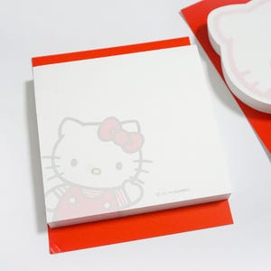 May include: A square white notepad featuring a light gray outline of the Hello Kitty character with a pink bow and red clothing. The bottom right corner of the notepad includes the text '©'76, '19 SANRIO'. A red paper sheet is visible underneath.