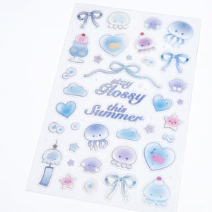 May include: A clear sticker sheet featuring various pastel-colored illustrations including jellyfish, bows, hearts, stars, clouds, and a dessert. The text "stay Glossy this Summer" is printed in the center in a blue cursive font with a white outline.