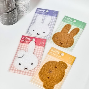 May include: Four Miffy character stickers in white, brown, and tan, each packaged on a patterned backing. The stickers feature the iconic bunny and bear designs with embroidered details. Rolls of matching decorative tape are visible in the background.