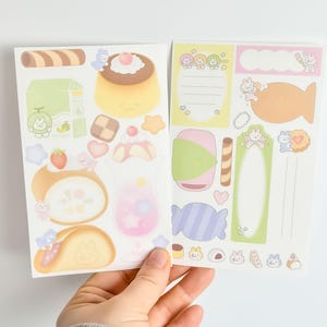 May include: Two sheets of colorful stickers featuring cute illustrations of desserts and snacks. The stickers include images of pudding, a parfait, a roll cake, and a candy. The stickers are held by a hand.