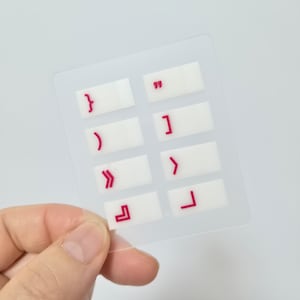 May include: A clear, rectangular sticker sheet with eight white rectangles, each featuring a red symbol. Symbols include parentheses, brackets, and quotation marks. The sticker sheet is held in a hand against a white background.