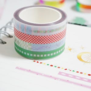 May include: A roll of decorative washi tape with various patterns, including red gingham, green borders, and pastel designs. The tape is stacked on a white surface, with additional patterned tape strips visible.