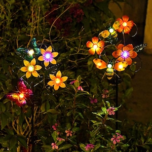 Pode incluir: Dois espetos solares de jardim com flores, borboletas e abelhas de metal iluminadas. Os ornamentos exibem pétalas vibrantes em laranja, amarelo e roxo com centros brilhantes, montados em hastes finas de metal preto entre a folhagem.