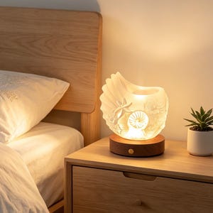 May include: A seashell-shaped lamp with intricate details, resting on a wooden base. The lamp emits a warm glow, illuminating the carved design of starfish and other sea creatures. The lamp sits on a wooden nightstand next to a small potted plant.