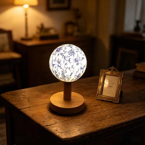 May include: A decorative table lamp with a spherical shade featuring a blue floral pattern on a white background. The lamp sits on a wooden base and stem, creating a warm, inviting glow. The lamp is on a wooden table.