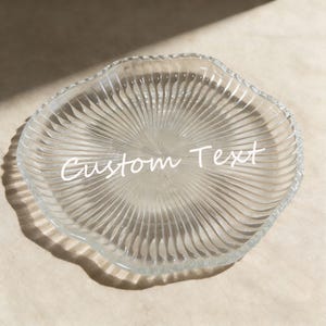 May include: A clear glass plate with a wavy, textured rim and a radial pattern. The plate has the words "Custom Text" written across the center in a white script font. The plate is on a light-colored surface.