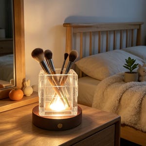 May include: A lit makeup brush holder with a wooden base. The clear, geometric-patterned container holds several makeup brushes. The warm light illuminates the brushes and the wooden base, creating a soft glow. The holder sits on a wooden bedside table.