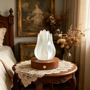 May include: A white, tulip-shaped lamp sits atop a round, wooden base with a gold-colored button. The lamp is on a small, antique wooden table with a lace doily. The background includes a bed and framed artwork.
