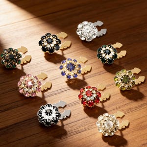May include: A collection of decorative flower-shaped jewelry pieces with various colored gemstones. Each piece has a gold-colored base and a C-shaped clasp. Gemstone colors include clear, black, green, pink, blue, red, and yellow.